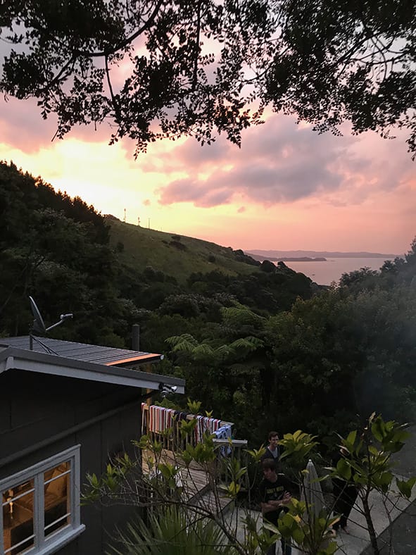 Photo looking down over a bach during sunset, with hills and the harbour in the background.